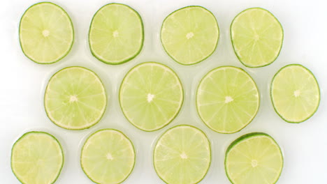 A-lot-of-lime-slices-lie-on-white-in-the-background-in-slow-motion-falling-water-splashes.